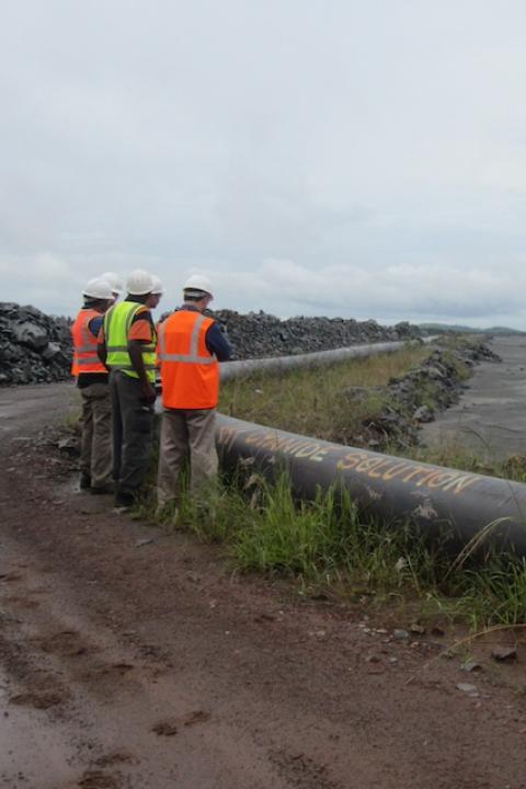 Workers stand next to a pipe labeled "cyanide solution" at a mine. 